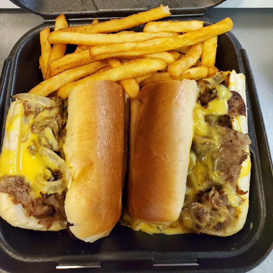 Two Romano's Deli Pizzeria cheesesteak sandwiches with fries in a black takeout container.